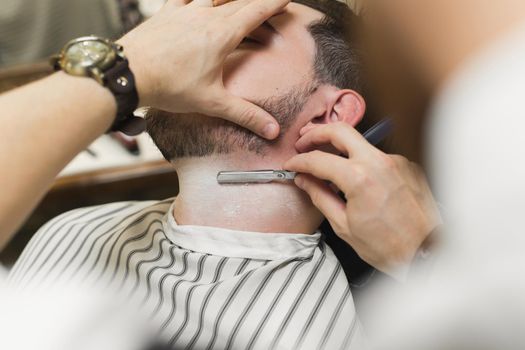 A young man is sitting in a chair in a barber shop while a barber shaves his beard. Preparation of the groom on the wedding day