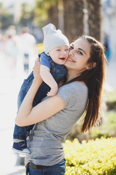 Cute stylish young mother lady kissing cheek nicely dressed son and enjoying nice sunny day. Son on mother hands and smiling.