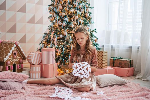 Adorable little girl sitting near the tree and making paper snow-flakes. Room decorated.