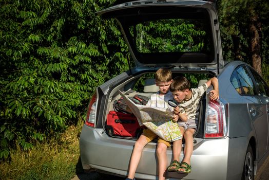 Two cute boys sitting in a car trunk before going on vacations with their parents. Kids sitting in a car examining a map. Summer break at school. Family travel by car