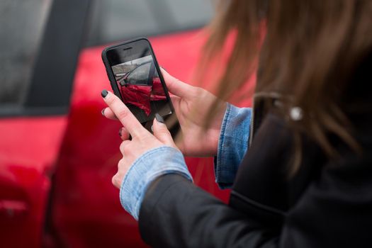 Woman using a smart phone to take a photo of the damage to her car caused by a car crash.