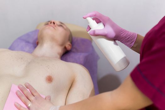 Close-up of mens Breasts in the beauty salon for the procedure of sugar hair removal. Beautician sprays disinfectant solution on the man's chest. Sugaring.
