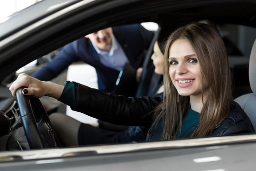 Side view of young beautiful woman sitting inside car and holding hand on steering wheel. She smiling and talking with manager of car dealership. Car agent representing inside of automobile