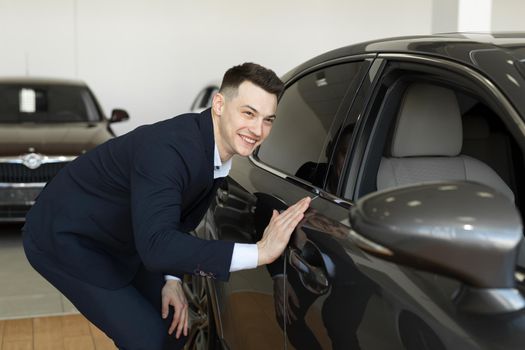Handsome young businessman in classic blue suit is smiling while examining car in a motor show.