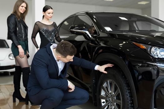 Attractive elegant man examining wheels of a new automobile on sale at dealership. Handsome male driver choosing new car to buy.