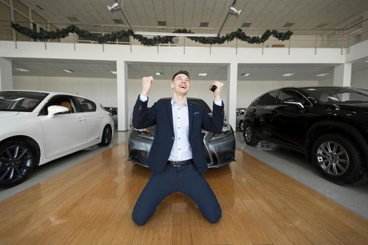 Young man in a suit is on his knees in a car dealership and is happy to buy a new car