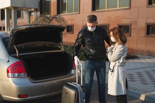Man and woman in protective medical masks and gloves with a suitcase leave the house by car during the quarantine and self-isolation. The coronavirus. Covid 19