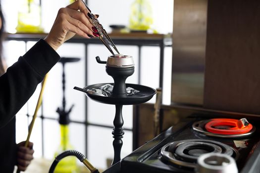 Women's hands hold hookah tongs and adjust the hot coals in a metal bowl. Black hookah stands in a restaurant or bar