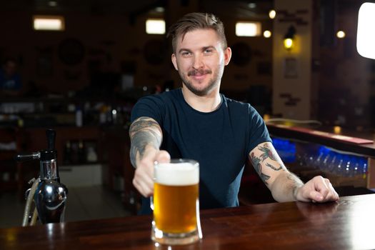 Handsome young male bartender in stretching out glass with beer and smiling while standing at the bar counter.