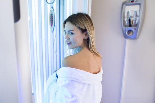 Sexy girl in a white robe is preparing for the tanning procedure in the Solarium. Vertical Solarium. Portrait of a slender girl in a modern Solarium.