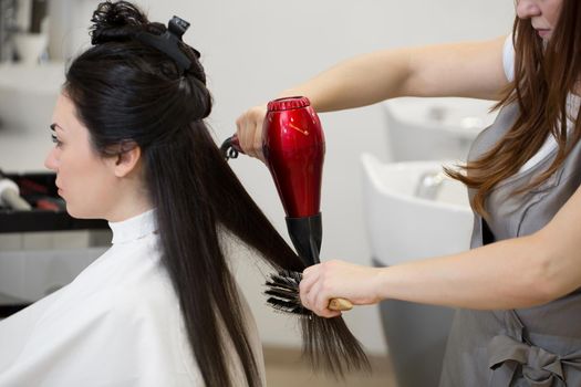 Hair stylist work on woman hairstyle in salon. Drying long brown hair with hair dryer and round brush.