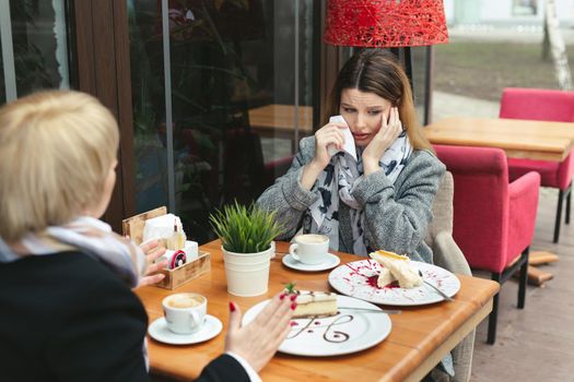 Sad and unhappy daughter sitting in cafe or restaurant with her mother, crying and wipes her tears with a paper napkin
