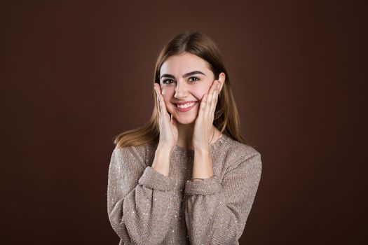 Pleasantly surprised woman looking with bugged eyes holding hands on cheeks opening her mouth in amazement not expecting to recieve such great present. Happy model isolated over brown background.
