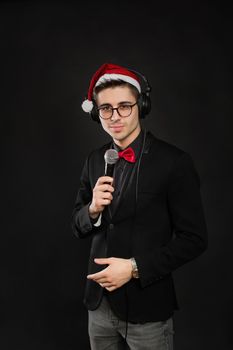 Portrait of a young male showman in glasses and a Santa Claus hat and headphones. With a microphone in his hands at a new year's party