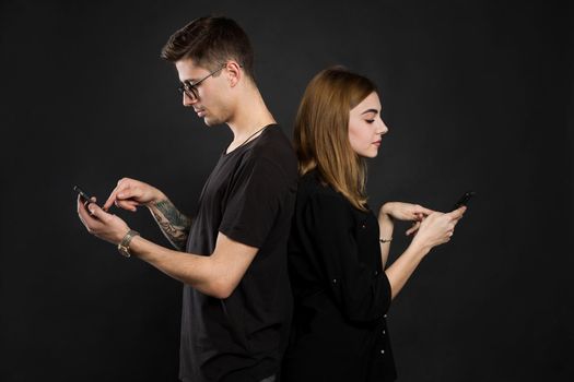 Profile portrait of young married couple, browsing information at their pdas, standing back to back, wearing casual outfits on black background. Another life in social nets.