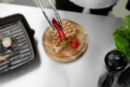 Close-up of the chef's hand as he puts chili peppers from the grill on a plate with a pair of tongs