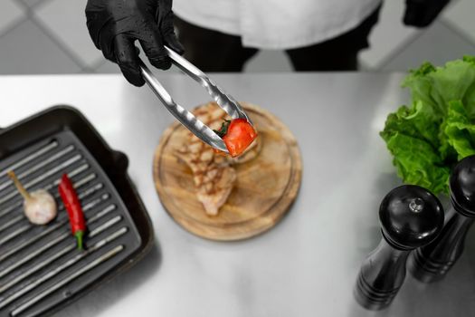 Close-up of the chef's hand as he puts a tomato from the grill on a plate with tongs