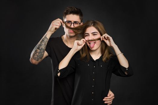 Beautiful young loving couple making fake moustache from hair while standing against black background. Funny moustache