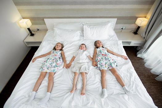 Twin sisters and a brother lie on a bed in a hotel room.