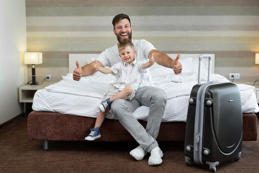 A young father and son sit on a bed in a hotel with a suitcase and laugh