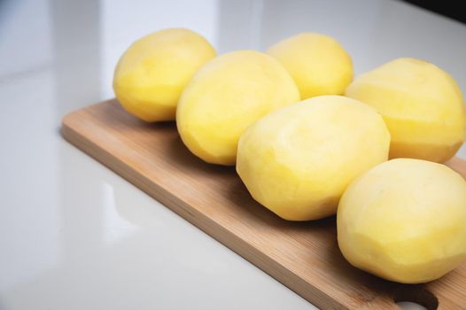Peeled fresh potatoes on a wooden cutting board on a white table.