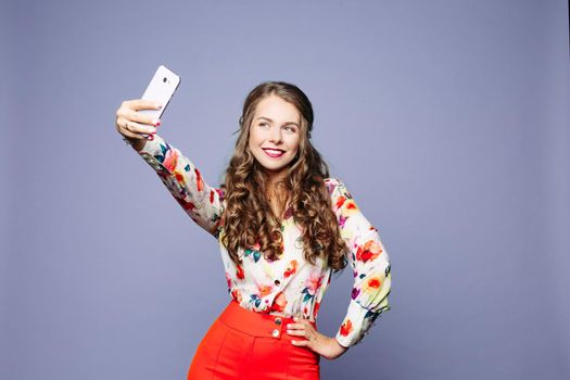 Attractive woman in floral shirt and red shorts taking self-portrait over violet background.