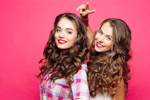 Two pretty happy girls with curly hair and, make up after beauty salon, smiling and showing peace by fingers. Stylish positivity women posing at pink studio. Emotionaly mood.