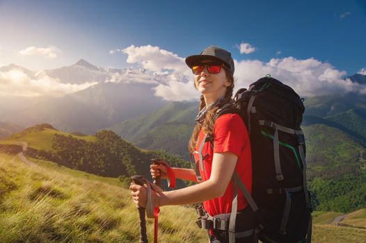 Caucasian traveler in a cap and sunglasses with a backpack on her back and trekking poles against the backdrop of high mountains and low clouds. Smiling and looking at the camera