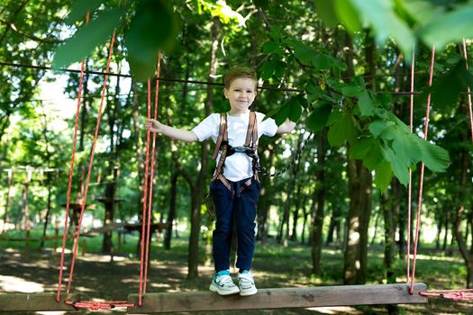 Small boy in climbing gear is walking along a rope road in an adventure Park, holding on to a rope and a carabiner