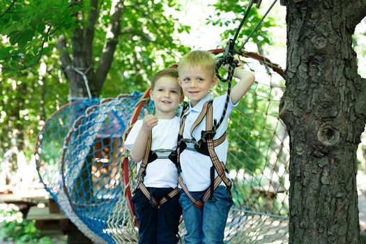 Two cute boys in an adventure park are doing rock climbing or passing obstacles on a rope road, hugging and giving a thumbs up