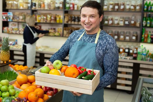 Smiling male salesman holds a wooden box with vegetables and fruits in the store