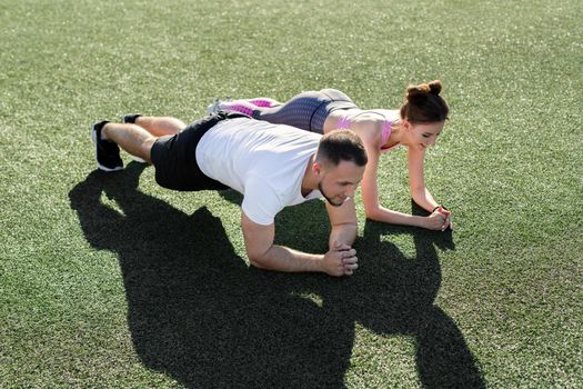 Close-up of a man and a woman doing a plank exercise on the grass at a sports stadium at sunset.