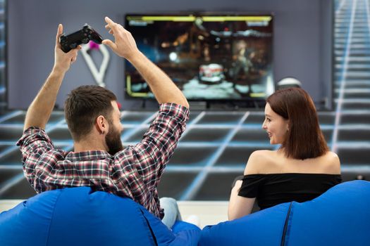 Young couple playing video games, sitting on chairs in a gaming club with controllers in their hands, a rear view from the TV screen