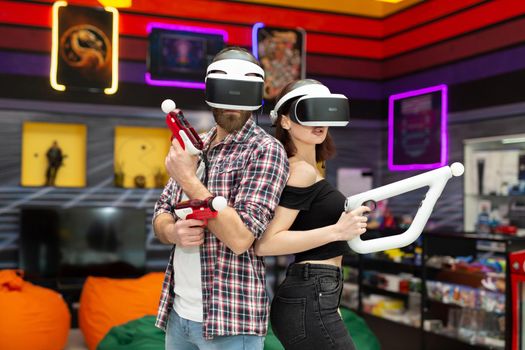 Friends, a man and a woman use a virtual reality headset with glasses and hand motion controllers and weapons in the play area