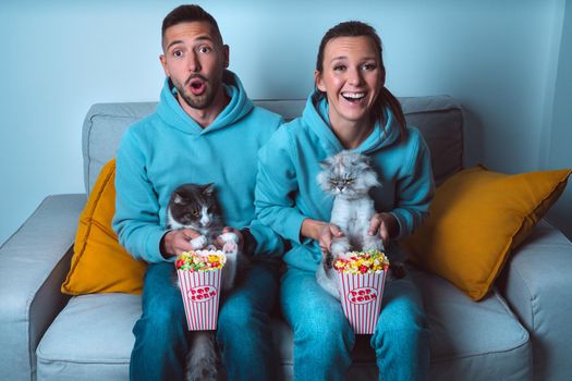 Young happy couple with popcorn and funny cats sitting on sofa watching comedy movie and enjoying time together at home