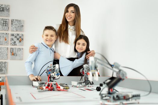 Young beautiful mother with her son and daughter pose at the school of robotics with robots from the constructor