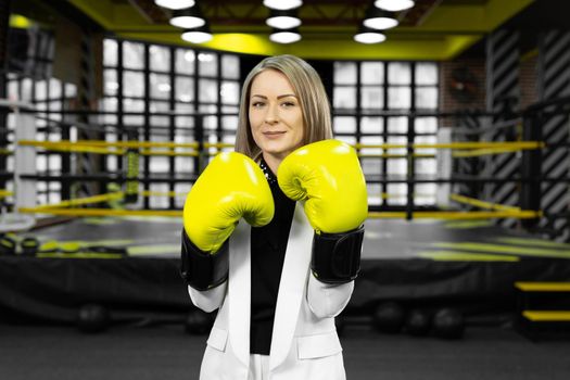 Determined, stylish businesswoman in yellow boxing gloves throws a punch at the camera against the backdrop of a boxing ring