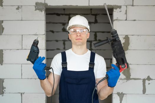 Professional construction worker in a white hard hat is holding a drill and a screwdriver