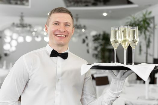 Smiling male waiter with glasses of white wine on a tray in a restaurant