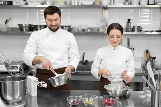 Pastry chef a man and a woman in a professional kitchen prepare a sponge cake, mix the ingredients and sift the flour