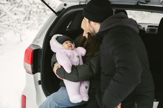 Young, happy family: a man, a woman and a baby are sitting in the trunk of a car in a winter snow-covered forest