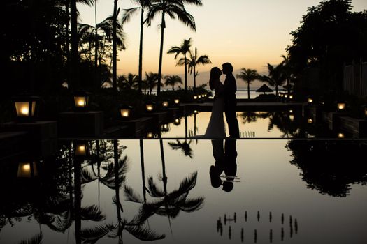 Silhouettes of the bride and groom at sunset. The reflection in the pool.
