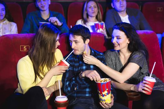 Two young girls and a guy watching a comedy in a cinema. Young friends watching movie in cinema. Group of people in theater with popcorn and drinks.