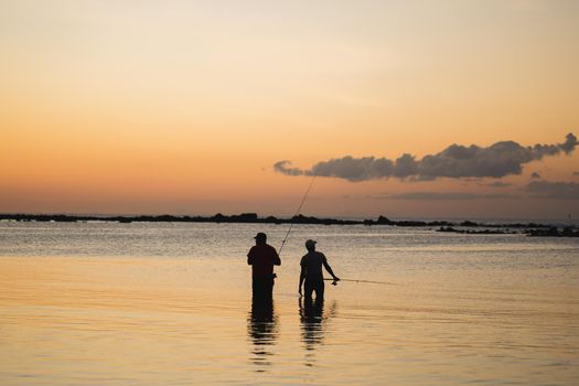 Two men fishing in the ocean from the beach at sunset.