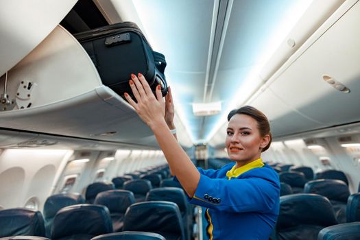 Woman flight attendant looking at camera and smiling while placing travel suitcase in overhead baggage locker in airplane passenger salon