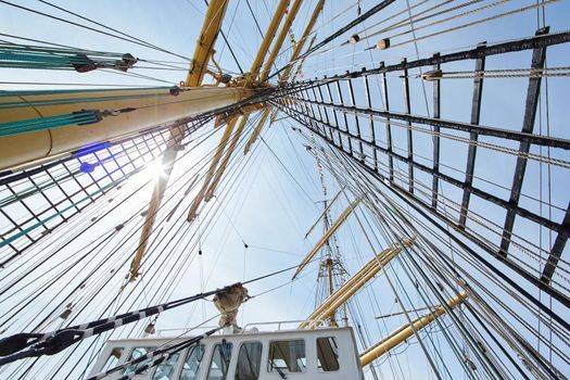 a view from below of the huge mast of an old sailing ship, many ropes hanging down from above, blue sky in the background on a sunny day