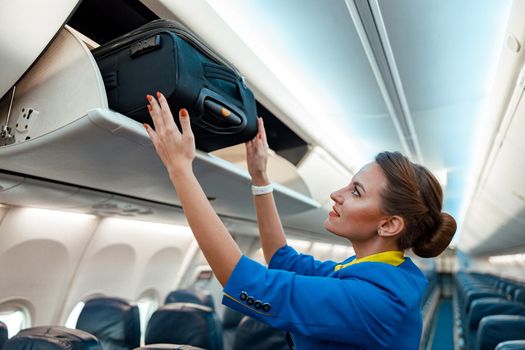 Woman flight attendant or air hostess placing travel bag in overhead baggage locker while standing in airplane passenger salon