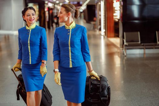 Two cheerful women stewardesses in air hostess uniform walking down airport terminal