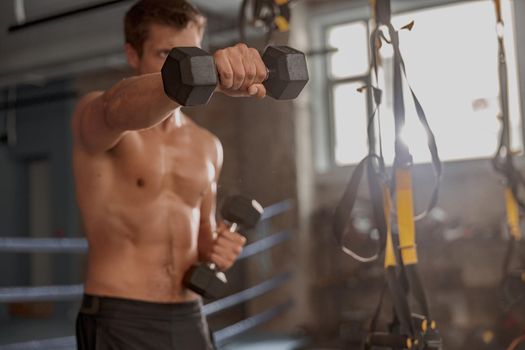 Confident male fitness instructor with athletic body holding pair of dumbbells in hands and exercising in gym