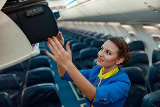 Woman stewardess flight attendant placing travel bag in overhead baggage locker while standing in aircraft passenger salon